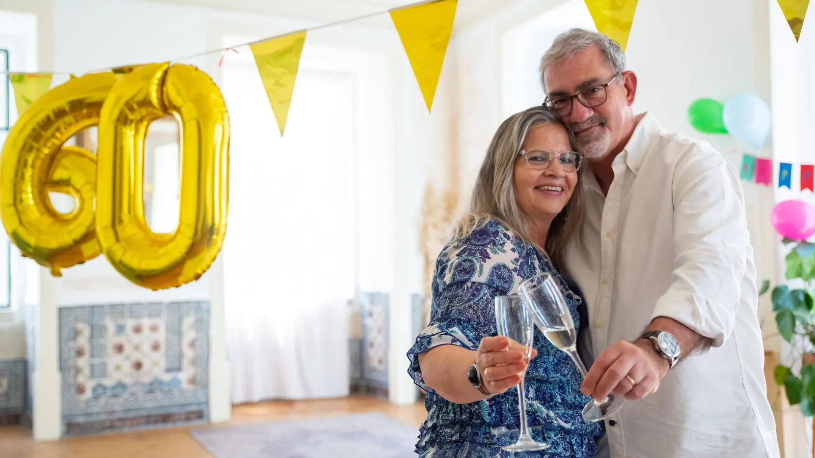 Smiling couple in their 60s raising glasses at a home celebration with gold balloons and bunting