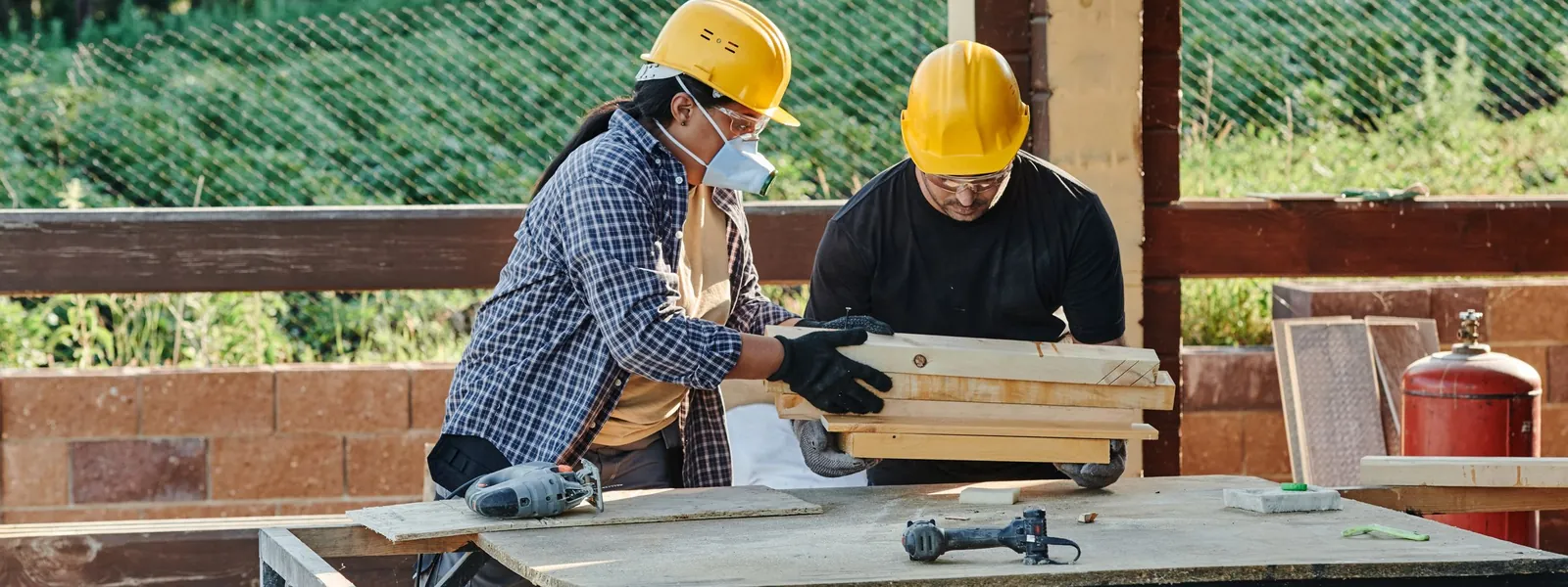 Two workers in hard hats measuring wood planks on a workbench at an outdoor job site