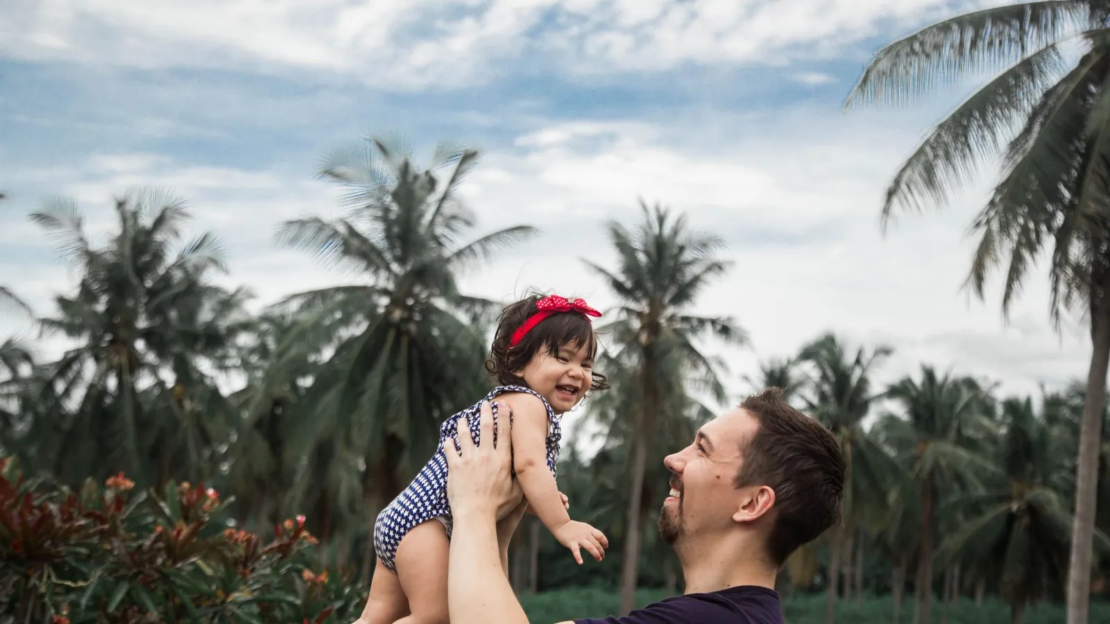 Father lifting his smiling baby daughter into the air outdoors under palm trees