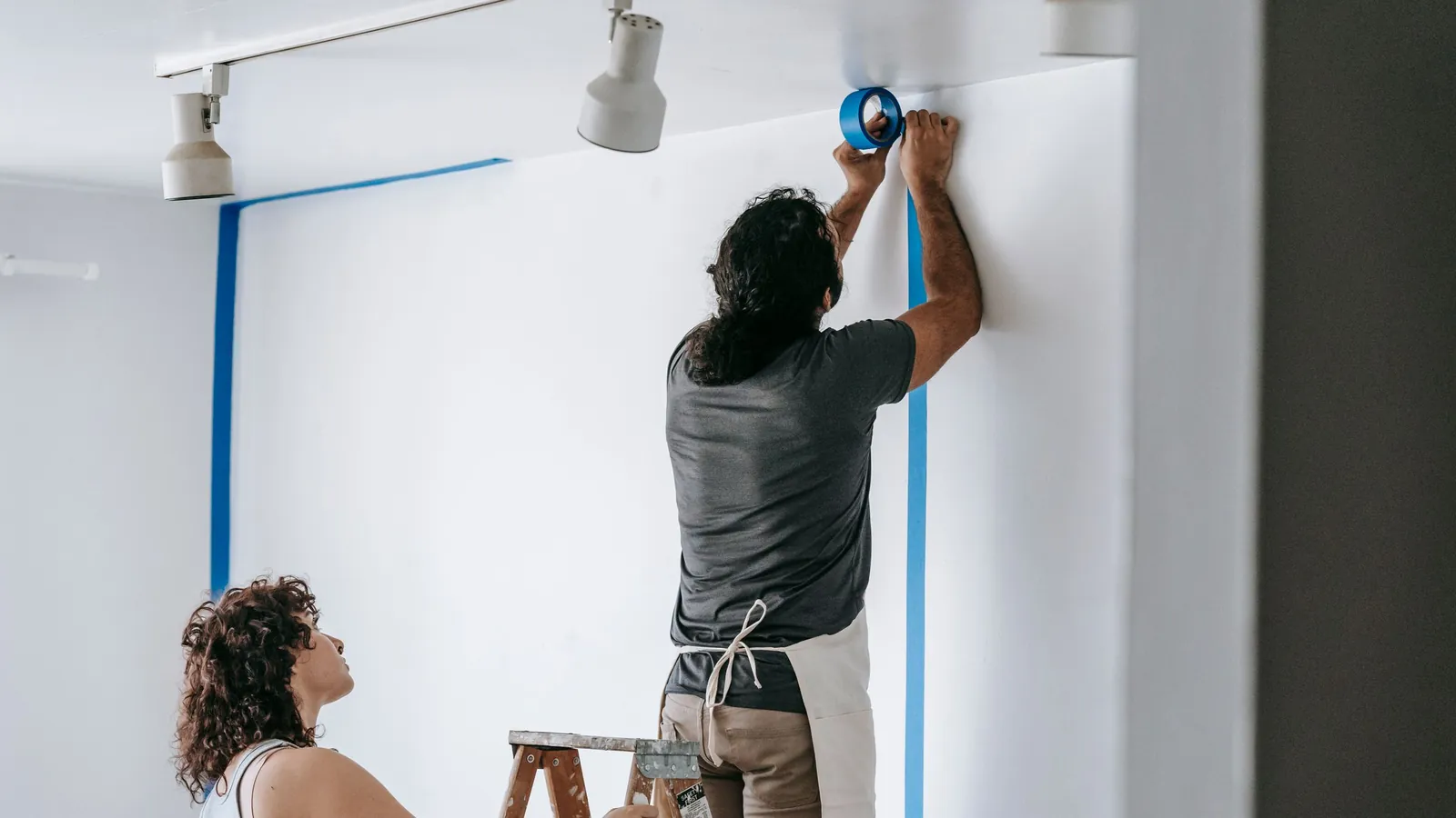 Painter taping a wall edge while a helper steadies the ladder