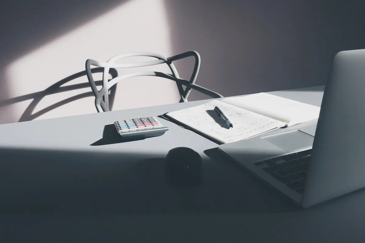 A desk with a laptop, a calculator, and an open notebook with a pen, in soft window light.