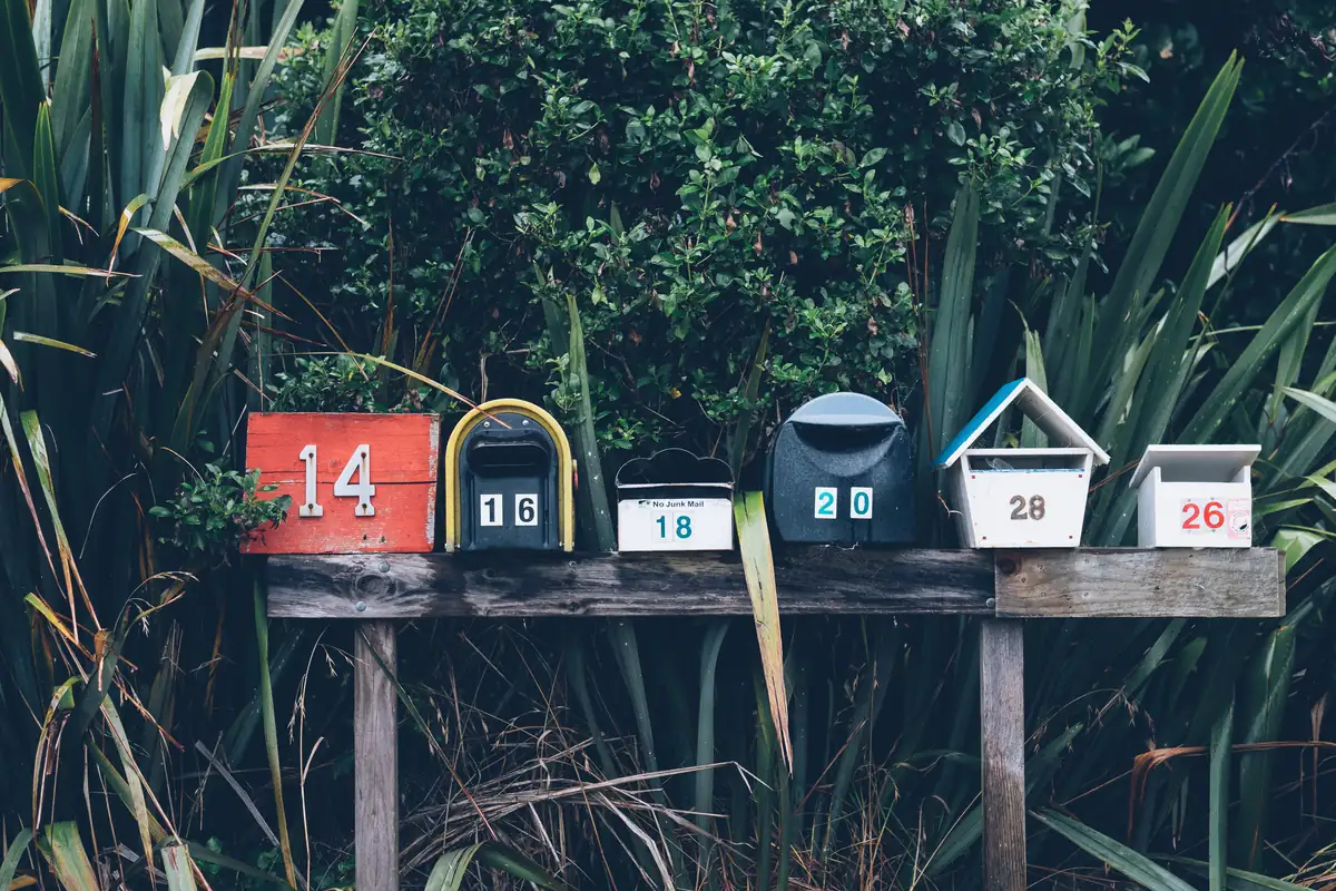 A row of six numbered mailboxes on a wooden stand in front of green plants.