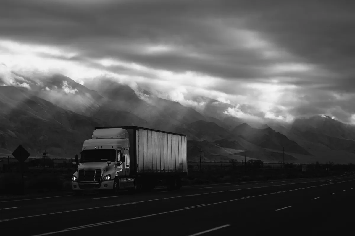 A black-and-white photo of a semi truck on a highway under storm clouds and mountains.