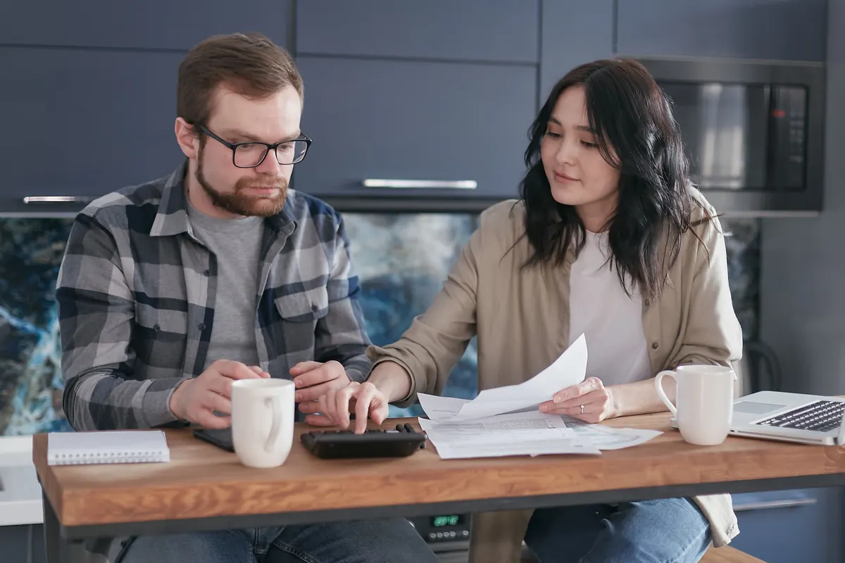 A couple at a kitchen table going over papers together, with a calculator and coffee mugs.