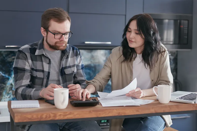 A couple at a kitchen table going over papers together, with a calculator and coffee mugs.