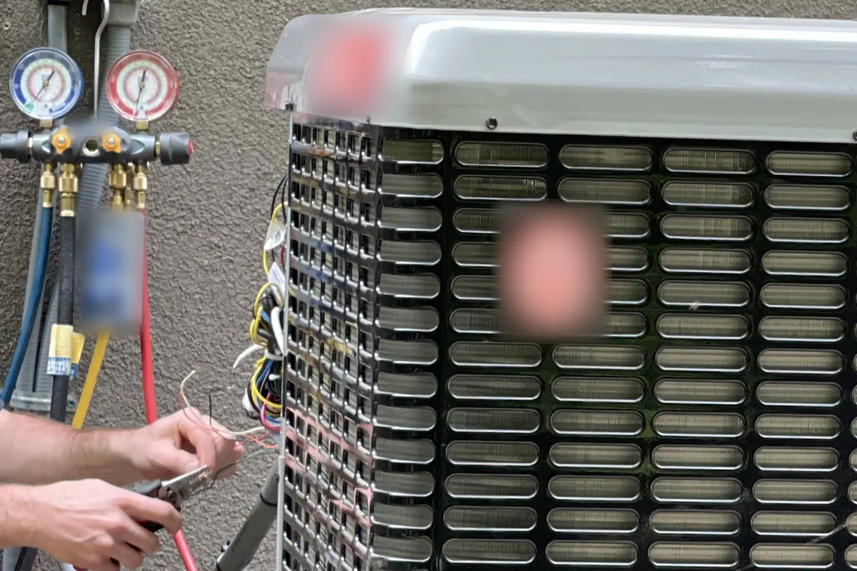 An HVAC technician's hands work on the wiring of a new outdoor heat pump unit, with gauges and hoses nearby.