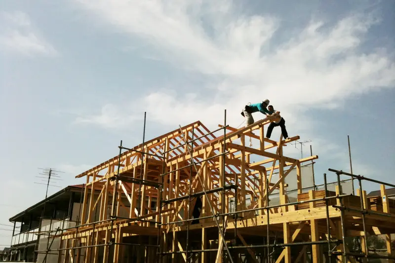 Two workers on top of a wooden house frame with scaffolding, against a blue sky.