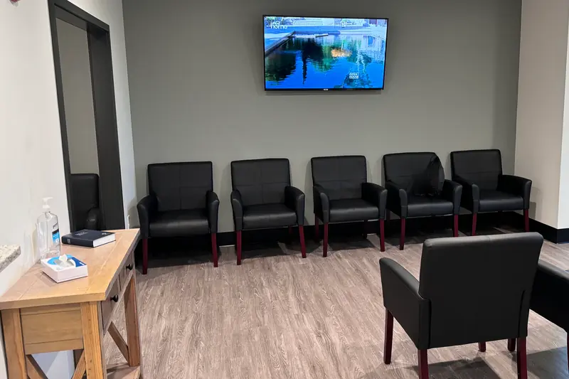 An empty doctor's office waiting room with rows of black chairs and a TV on the wall.