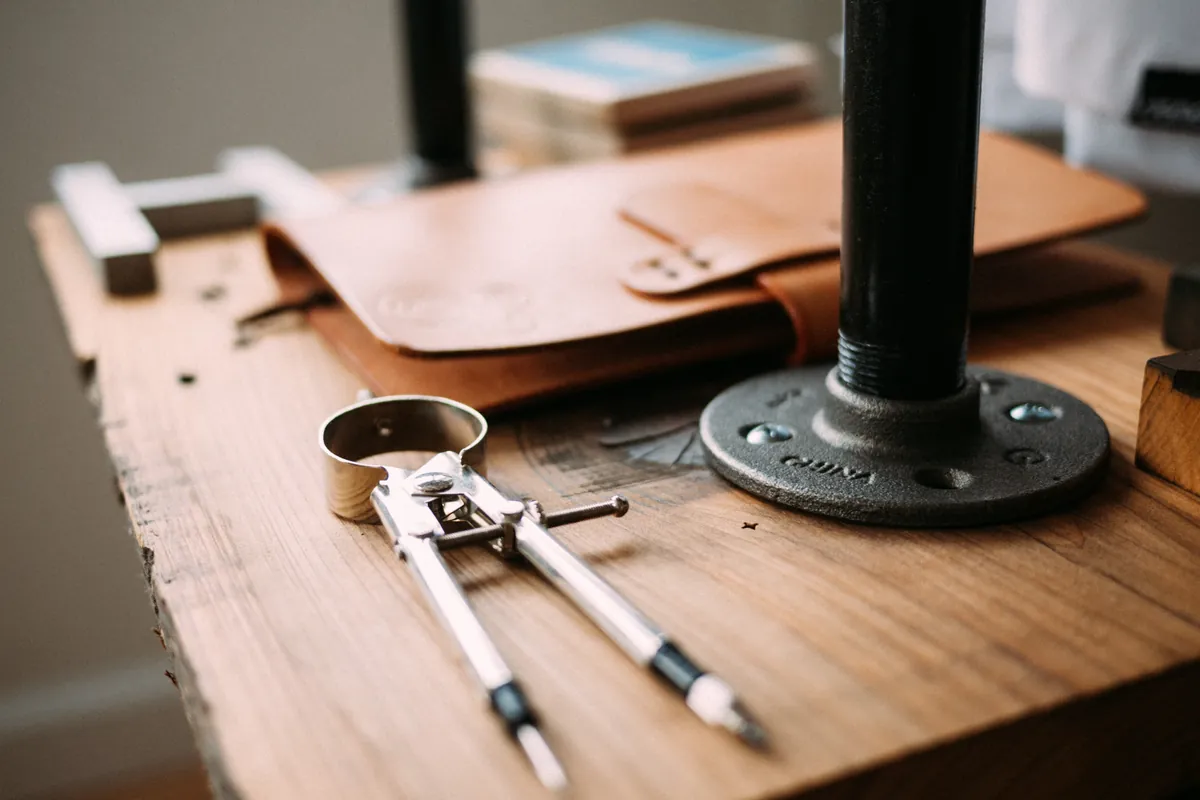 A wooden workbench with a drafting compass, a leather folio, and a metal pipe stand.