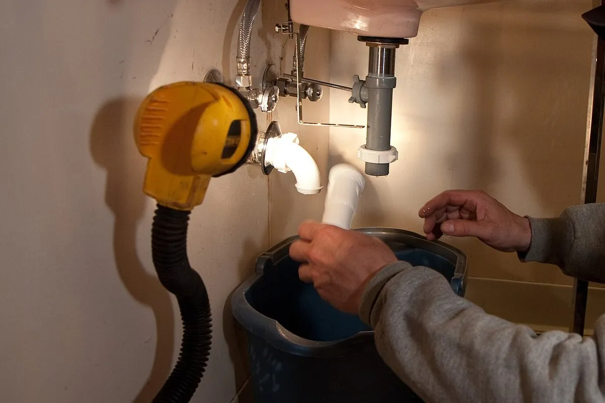 Hands working on the drain pipe under a bathroom sink, with a bucket below.