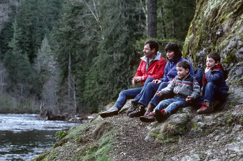 A family with two young boys resting on a rocky trail beside a river in a forest.